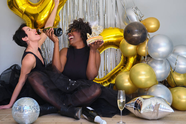 Dvě multiiraciální ženy na 40 narozeninové oslavě se smíchem pijící šampaňské z láhve. - Fotografie, Obrázek