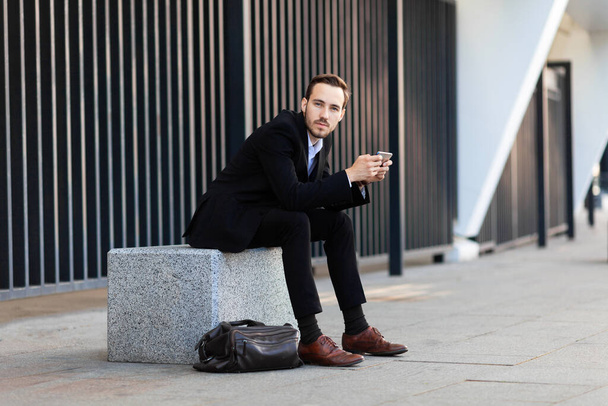Young businessman in suit sits and plays on phone. Social networks addiction. Using mobile application. - Photo, Image