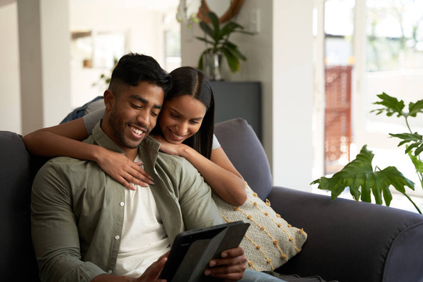 Happy young biracial couple wearing casual clothing embracing over sofa with digital tablet at home - Fotografie, Obrázek