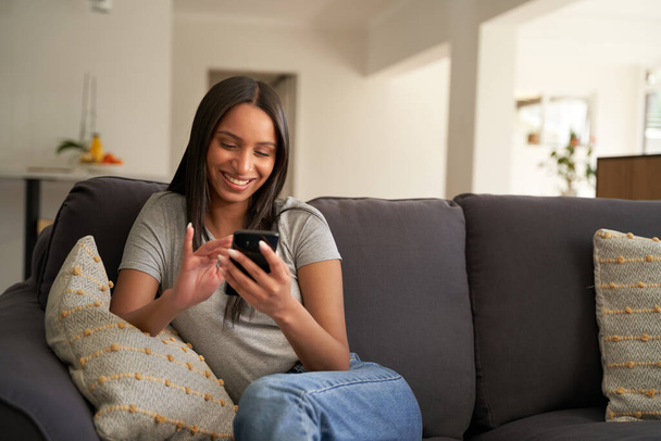 Mujer biracial joven usando ropa casual usando teléfono móvil mientras está sentada en el sofá en casa - Foto, Imagen
