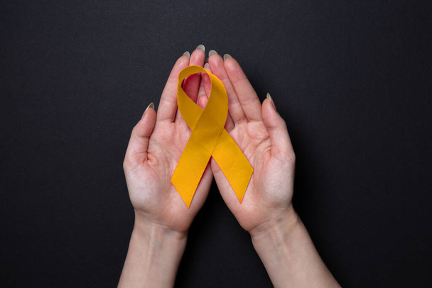 Female's hands hold a golden ribbon in their palms. Black background. Copy space. Flat lay. The concept of childhood cancer. - 写真・画像