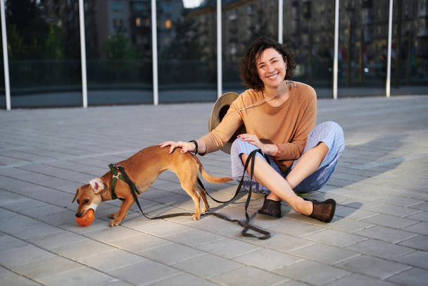 Jeune femme adulte dans la ville avec un chien assis et étreignant, souriant. Caresser le chien, nourrir, s'amuser Photo de haute qualité - Photo, image