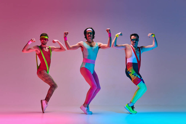 Sport show. Three funny men in colorful sportswear doing aerobics exercises against gradient blue pink studio background in neon light. Concept of sportive and active lifestyle, humor, retro style. Ad - 写真・画像