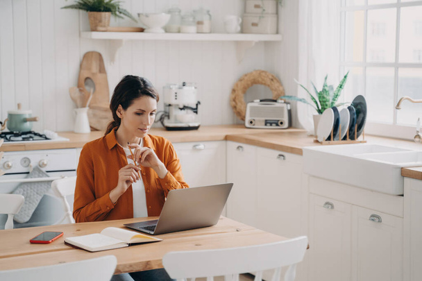 A elfoglalt nő otthon dolgozik, a konyhaasztalnál ül laptoppal. Töprengő háziasszony üzletek online, tervez vásárlások, teszi a tennivalók listáját, nyelvtanulást - Fotó, kép