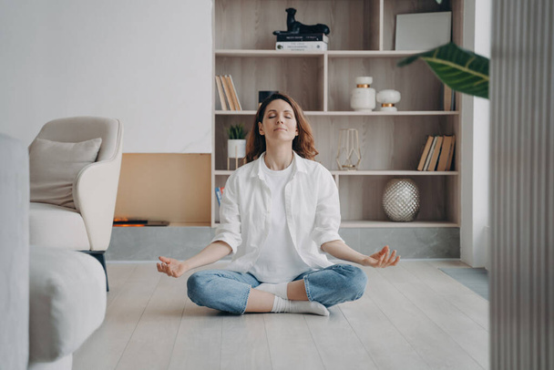 Donna calma in posizione di loto, meditando in soggiorno. Yoga, aria fresca, da solo a casa. Attività ricreative, benessere. - Foto, immagini