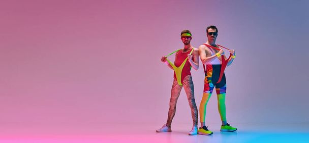 Stylish young men in colorful retro sportswear posing over gradient blue pink studio background in neon light. Concept of sportive and active lifestyle, humor, retro style. Banner. Copy space for ad - Фото, зображення