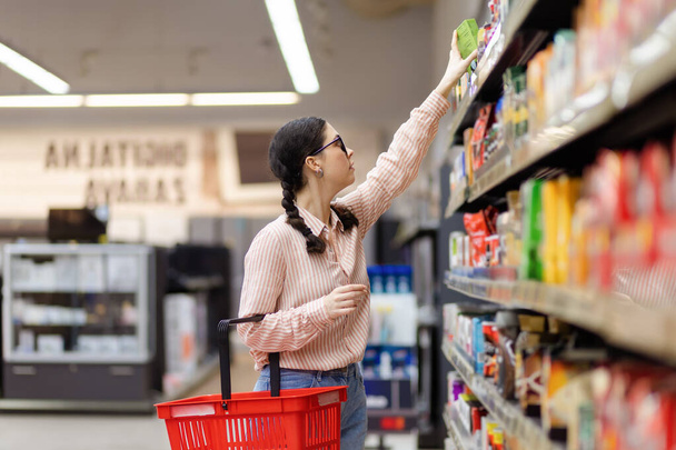 Visit grocery store. Side view of young pretty caucasian woman holds cart and reaches hand to top shelf. Concept of shopping and consumerism. - Fotografie, Obrázek