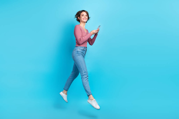 Full length profile photo of excited gorgeous person jumping hold smart phone isolated on blue color background. - Fotó, kép