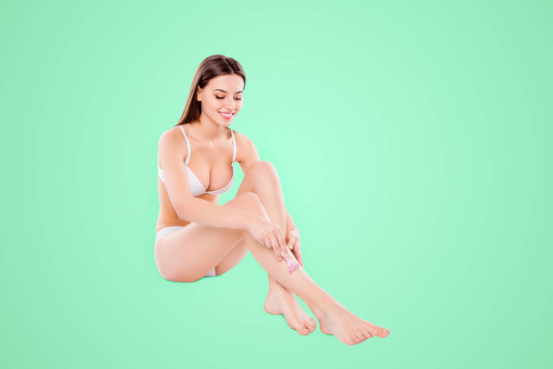 Portrait of pretty woman shaving slim long legs carefully sitting over white background. - 写真・画像