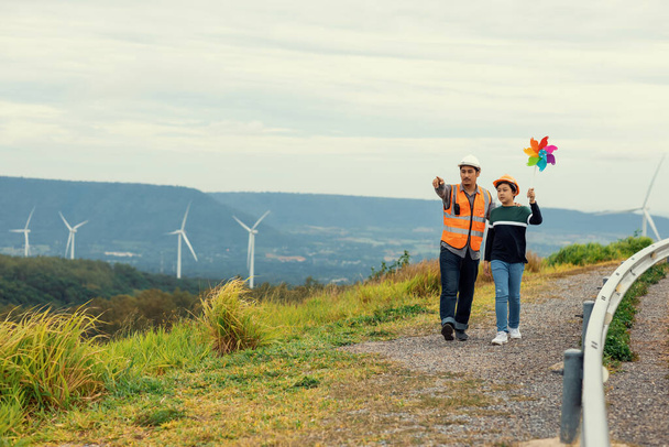 Engineer with his son holding windmill toy on a wind farm atop a hill or mountain. Progressive ideal for the future production of renewable, sustainable energy. Energy generated from wind turbine. - Foto, Bild
