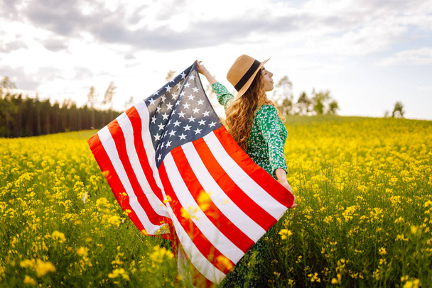 Young woman with american flag on blooming meadow. 4th of July. Independence Day. Patriotic holiday. USA flag fluttering in the wind. - Photo, image