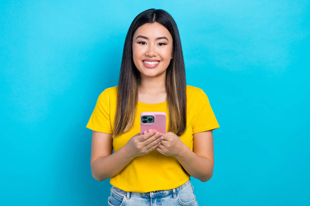 Photo of positive cheerful cute girl with straight hairdo dressed yellow t-shirt hold smartphone isolated on blue color background. - Foto, immagini