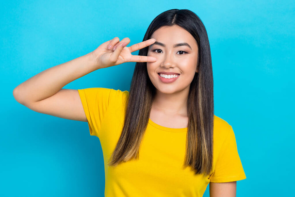 Portrait of cute pretty asian girl say hi show v sign gesture near eye wear yellow t-shirt isolated shine blue teal color background. - Zdjęcie, obraz
