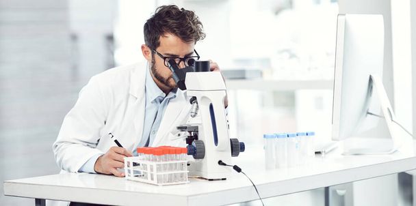 Robić to, co lekarze robią najlepiej. skupiony młody naukowiec robi notatki patrząc przez mikroskop i siedząc w laboratorium - Zdjęcie, obraz