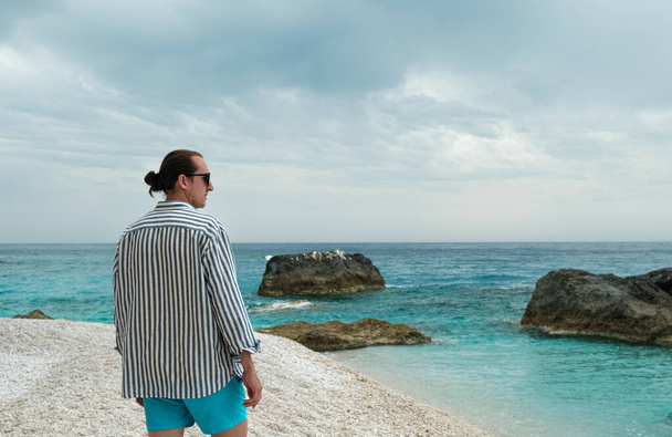 Giovane uomo in piedi sulla spiaggia con mare blu e rocce e cielo lunatico sullo sfondo - Foto, immagini