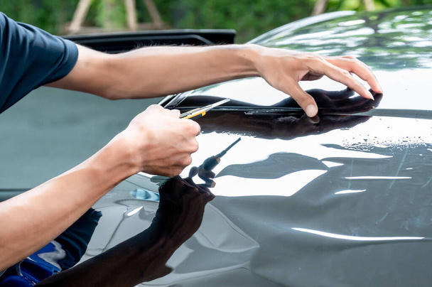 Παραπέτασμα παραθύρων αυτοκινήτων και εγκατάσταση χρωματισμού. Αρσενικό auto εξειδικευμένος εργάτης χέρι κρατώντας κόφτη απαλά προσεκτικά κοπής αυτοκίνητο εμπρός παρμπρίζ φιλμ στην επιφάνεια γυαλιού. - Φωτογραφία, εικόνα