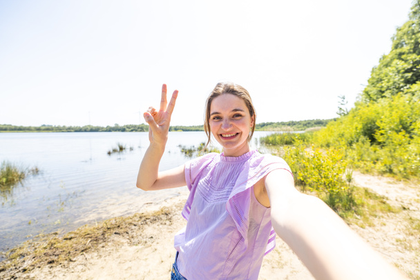 自然と外のスマートフォン機器で自撮り写真を撮る幸せな魅力的な女の子。湖の側で笑顔で楽しい美しい若い女性。女性が楽しむ健康的なライフスタイルのコンセプト - 写真・画像