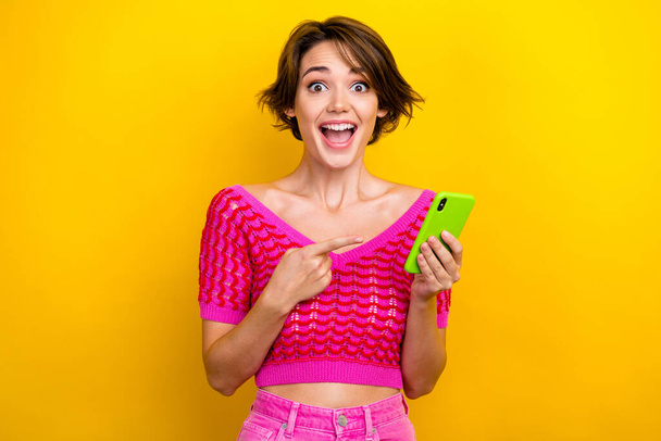 Retrato de mujer divertida loca extática con corte de pelo corto desgaste top de punto rosa dirigiendo en el teléfono inteligente aislado sobre fondo de color amarillo. - Foto, imagen