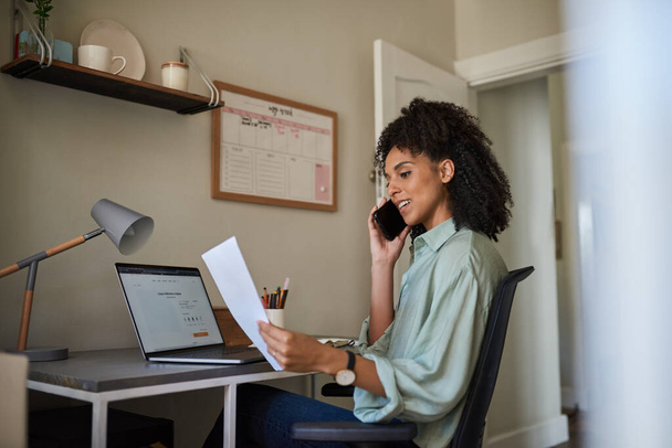 Young African female entrepreneur talking on a phone while reading paperwork and working on a laptop at a desk in her home office - Fotoğraf, Görsel