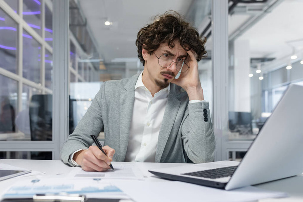 Upset businessman behind paper work inside modern office, mature man with beard reading financial reports and account documents unhappy with results and disappointed with achievements - Фото, зображення