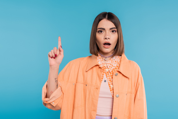summer fashion, shocked young woman with short hair and piercing in nose and tattoos posing in casual outfit on blue background, everyday makeup, orange shirt, generation z, showing and pointing up - Photo, Image