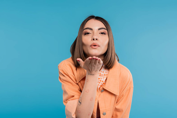 sending air kiss, brunette young woman with short hair, piercing in nose and tattoos posing in casual outfit on blue background, everyday makeup, orange shirt, generation z, blow kiss - Photo, Image
