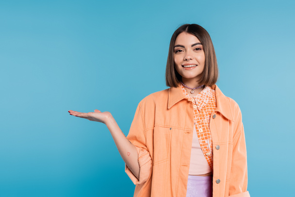 happy young woman with short hair, piercing in nose and tattoos demonstrating something in camera on blue background, showing, orange shirt, summer outfit generation z  - Photo, Image