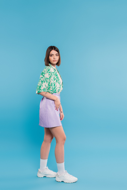 summer fashion, young brunette woman with short hair posing in shirt with palm tree print, skirt and white sneakers on blue background, carefree, tattooed, nose piercing, casual attire, full length  - 写真・画像