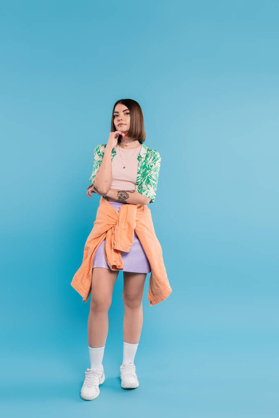 generation z, young brunette woman with short hair posing in shirt with palm tree print, skirt and white sneakers on blue background, tattooed, nose piercing, casual attire, full length  - Φωτογραφία, εικόνα