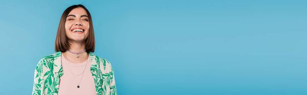 cheerful young woman with short brunette hair wearing shirt with palm tree print, smiling with closed eyes on blue background, casual attire, gen z fashion, emotional, happiness, banner  - Foto, imagen