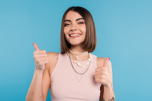 positivity, tattooed young woman with short brunette hair in tank top smiling and showing thumbs up on blue background, casual attire, gen z fashion, happiness, like gesture - Photo, Image