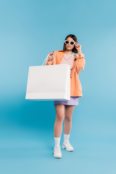 Einkaufsbummel, brünette junge Frau im Tank-Top, Rock, Sonnenbrille und orangefarbenem Hemd posiert mit Einkaufstasche auf blauem Hintergrund, lässige Kleidung, stilvolles Posen, moderne Mode  - Foto, Bild