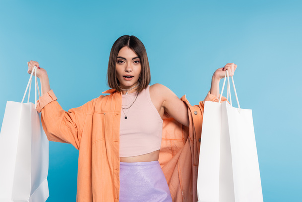 summer trends, pierced young woman in fashionable outfit posing with shopping bags on blue background, casual attire, stylish, generation z, modern fashion, orange shirt, tank top and skirt  - Foto, Bild