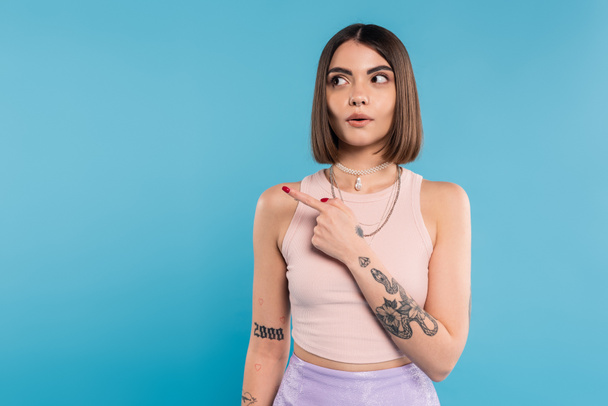 surprised woman showing something and looking away, stylish and tattooed model with nose piercing pointing with finger on blue background, generation z, emotional  - Photo, Image