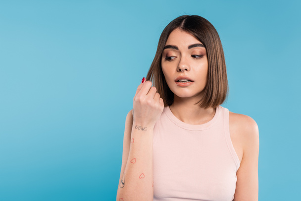 hair damage, stress, concerned brunette young woman touching her hair on blue background, tattooed, nose piercing, tank top, generation z, casual attire, thinning hair  - Photo, Image