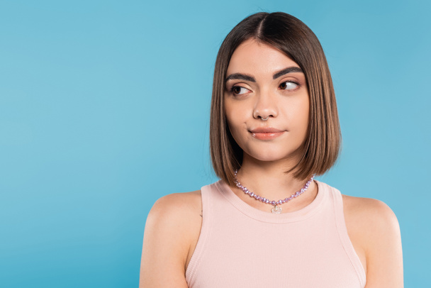 doubting, young brunette woman with short hair and nose piercing standing in tank top and looking away on blue background, youth culture, skepticism, generation z, summer fashion  - Fotoğraf, Görsel