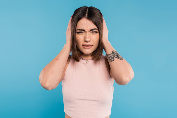 ημικρανία, τατουάζ νεαρή γυναίκα με σκουλαρίκι στη μύτη και κοντά μαλλιά αγγίζοντας το κεφάλι, ενώ πάσχουν από πονοκέφαλο σε μπλε φόντο, γενιά z, κούραση, άγχος  - Φωτογραφία, εικόνα