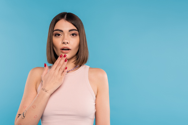 astonished, tattooed young woman with nose piercing and short hair holding hand near mouth on blue background, generation z, fashionable, trendy summer fashion  - Photo, Image