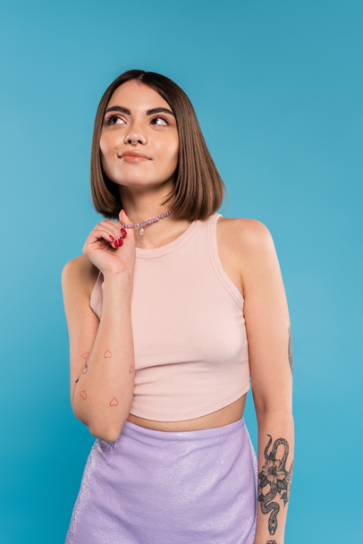 dreamy, tattooed young woman with nose piercing and short hair touching silver necklace and looking away on blue background, generation z, fashionable, trendy summer fashion  - Foto, imagen