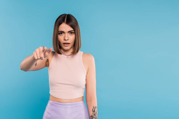 pointing at camera, emotional young woman with short hair, tattoos and pose piercing gesturing on blue background, generation z, displeased, casual attire, everyday makeup  - Φωτογραφία, εικόνα