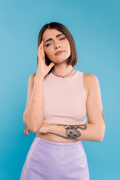 having headache, brunette young woman with short hair, tattoos and nose piercing touching head on blue background, generation z, displeased, casual attire, migraine, feeling pain - Photo, Image