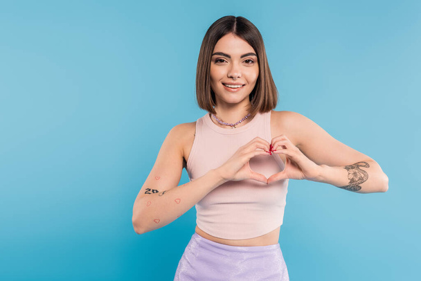 positivity and love, happy young woman with short hair, tattoos and nose piercing showing heart gesture with hands on blue background, generation z, cheerful, casual attire - Photo, Image