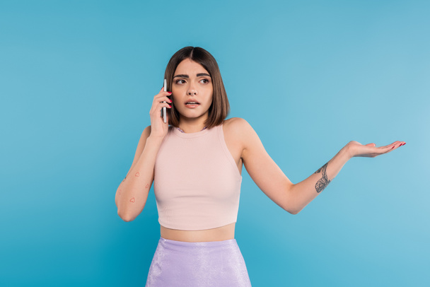 phone call, confused young woman with short hair, tattoos and nose piercing gesturing while talking on smartphone on blue background, casual attire, gen z fashion, personal style  - Foto, Imagen