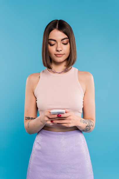 woman texting on smartphone, short hair, tattoos and nose piercing using mobile phone on blue background, casual attire, gen z fashion, personal style, everyday makeup  - Фото, зображення