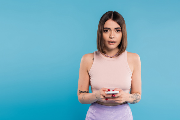 messaging on smartphone, shocked, young brunette woman short hair, tattoos and nose piercing using mobile phone on blue background, casual attire, gen z fashion, social media influencers  - Foto, afbeelding