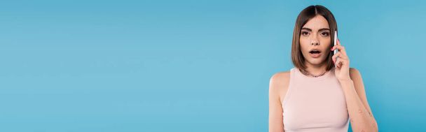 phone call, shocked young woman with short hair, tattoos and nose piercing talking on smartphone on blue background, casual attire, gen z fashion, opened mouth, emotional, banner - Photo, Image