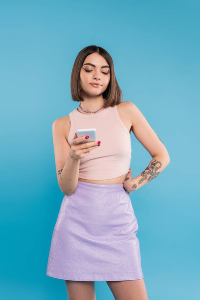 sending a message, attractive young woman short hair, tattoos and nose piercing using mobile phone on blue background, casual attire, gen z fashion, social media influencers  - Foto, immagini
