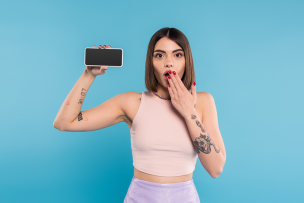 смартфон с чистым экраном, шокированная молодая женщина с короткими волосами, татуировки и пирсинг носа держа мобильный телефон на синем фоне, ген z мода, социальные медиа влиятельные, прикрыть рот  - Фото, изображение