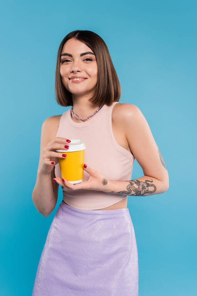 Kahve içmek için, kısa saçlı, dövmeli ve burnu piercing 'li mutlu genç bir kadın elinde mavi arka planda kağıt bardak, z nesli, yaz trendi, çekici, kahve kültürü, günlük tarz  - Fotoğraf, Görsel