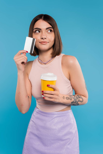 to go coffee, young woman with short hair, tattoos and nose piercing holding paper cup and credit card on blue background, generation z, summer trends, attractive, coffee culture, dream - Photo, Image